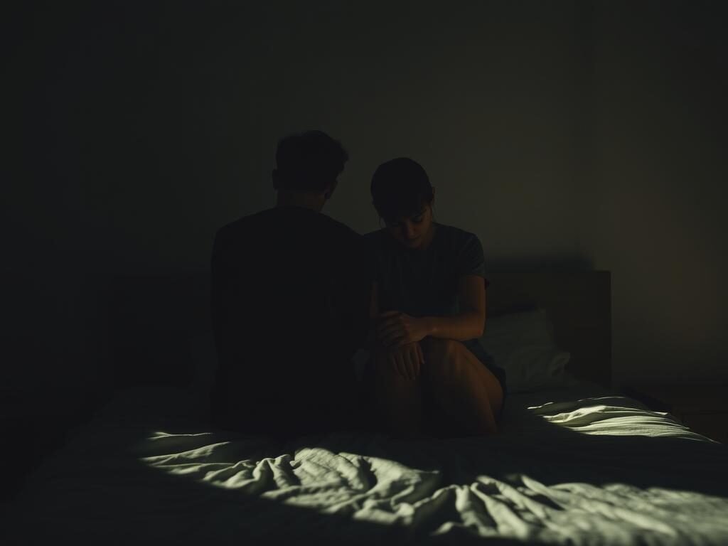 A dimly lit bedroom, the air heavy with tension. Two figures, their faces obscured, sit together on the edge of the bed, shoulders hunched, hands clasped tightly. The scene evokes a sense of unease, a disconnect between the physical proximity and the emotional distance. Subtle shadows cast across the scene, lending a moody, introspective atmosphere. The lighting is soft, almost melancholic, casting a warm glow that underscores the emotional weight of the moment. The composition is tight, focused on the two figures, emphasizing the isolation and the weight of their internal struggles. An unseen force seems to pull them apart, creating a tangible rift in their relationship.